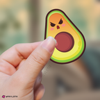 Hand holding a cartoon avocado sticker with an angry expression against a blurred background