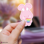 Hand holding a changbin skzoo sticker with a rainbow heart against a blurred background