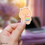 Hand holding a han skzoo sticker with a rainbow heart against a blurred background