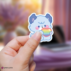 Hand holding a jooyeon xdinary monsters sticker with a rainbow heart against a blurred background