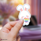 Hand holding a lee know skzoo sticker with a rainbow heart against a blurred background