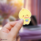 Hand holding a mingi aniteez sticker with a rainbow heart against a blurred background