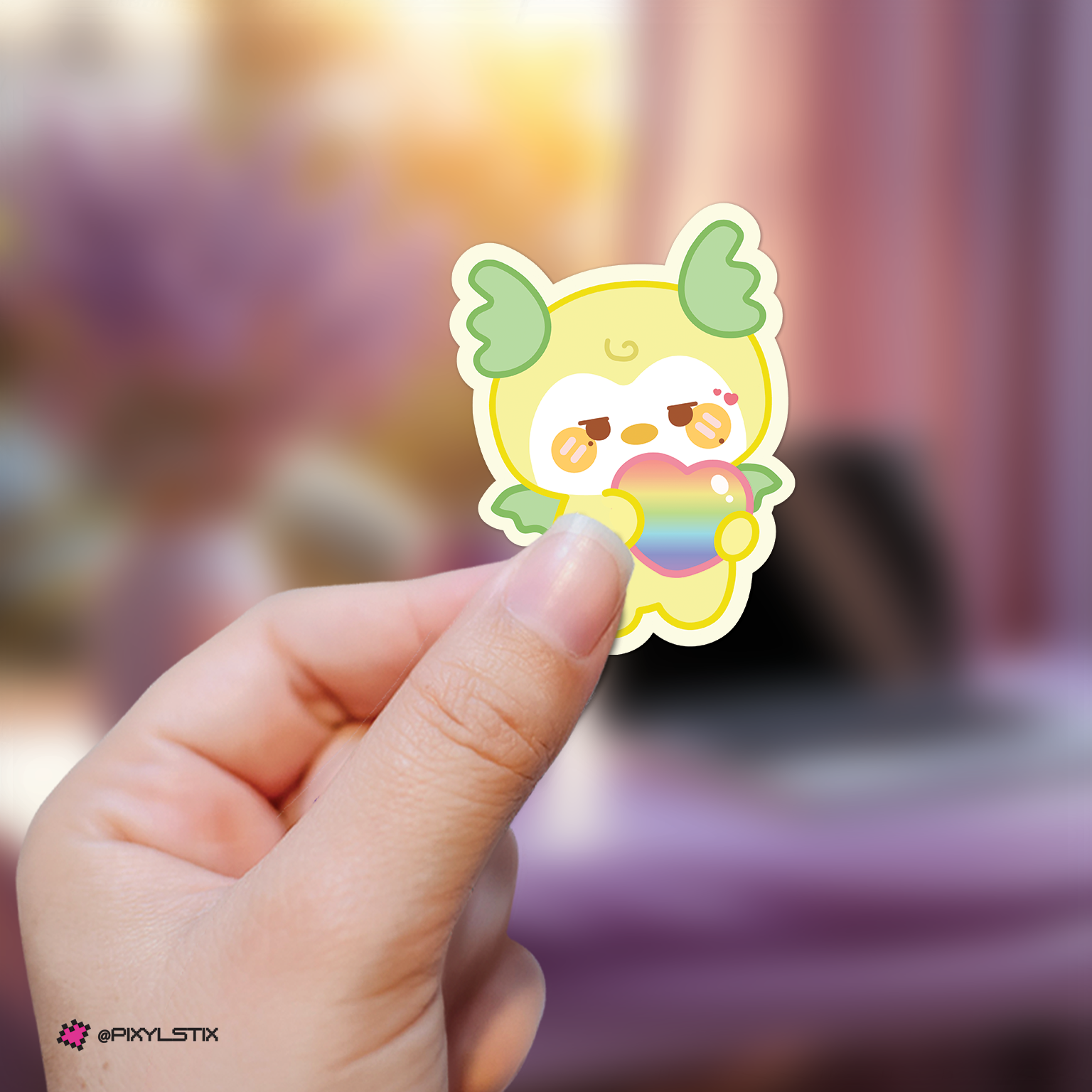 Hand holding a mingi mighteez sticker with a rainbow heart against a blurred background