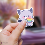 Hand holding a o.de xdinary monsters sticker with a rainbow heart against a blurred background