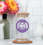 Clear glass tumbler with wooden lid and straw, featuring a purple decal, on a white surface with flowers in the background.