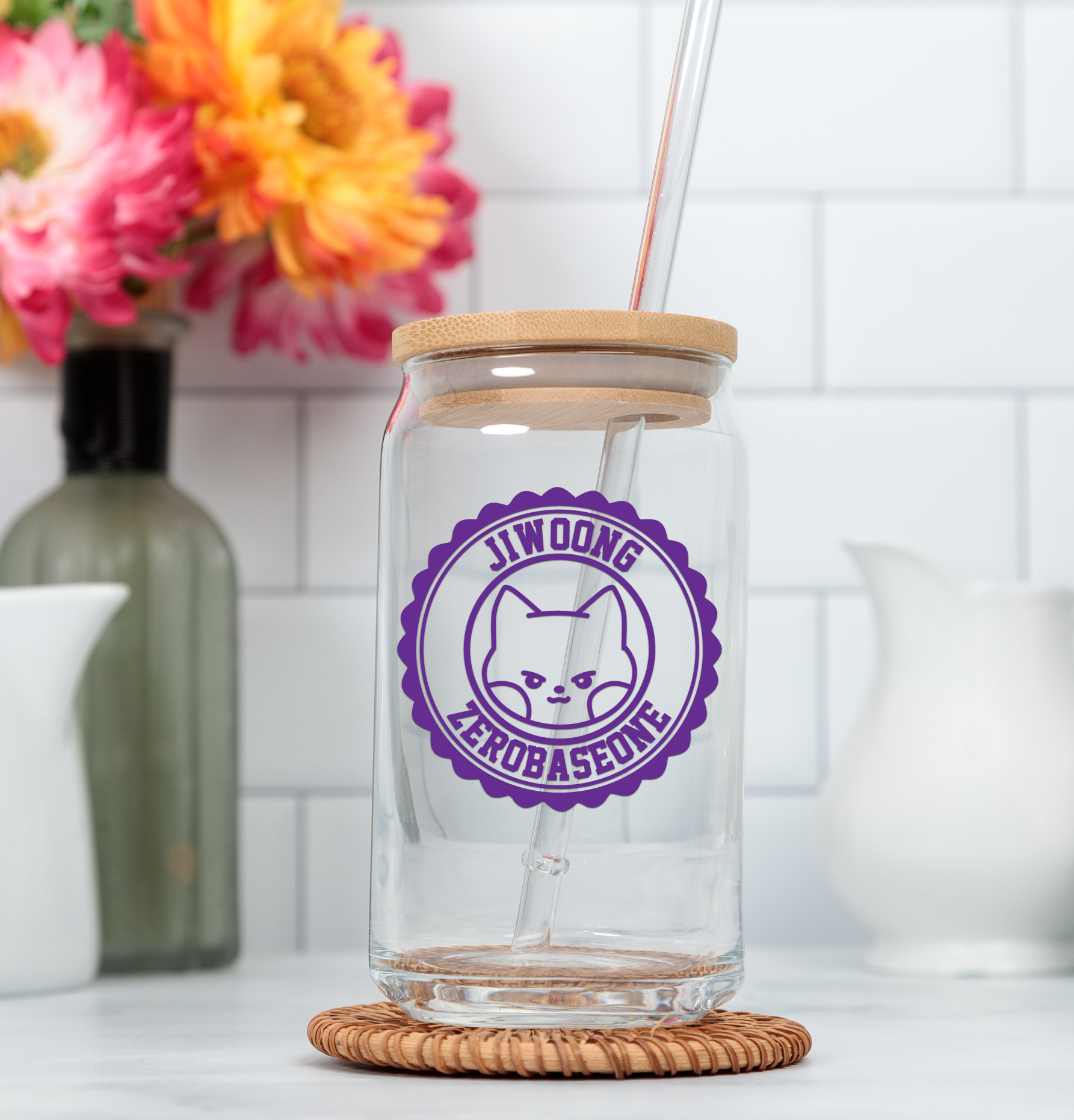 Clear glass tumbler with wooden lid and straw, featuring a purple decal, on a white surface with flowers in the background.