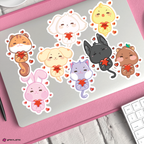 A set of cute aniteezz stickers on a silver laptop, surrounded by a pink textured mat with a white keyboard visible in the corner.