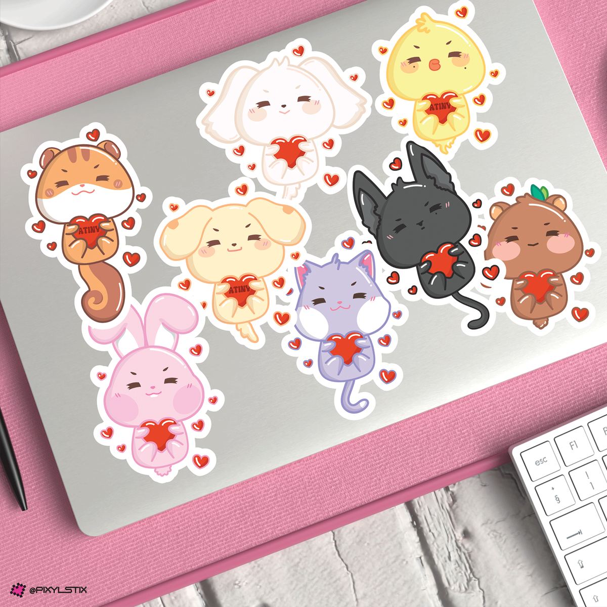 A set of cute aniteezz stickers on a silver laptop, surrounded by a pink textured mat with a white keyboard visible in the corner.