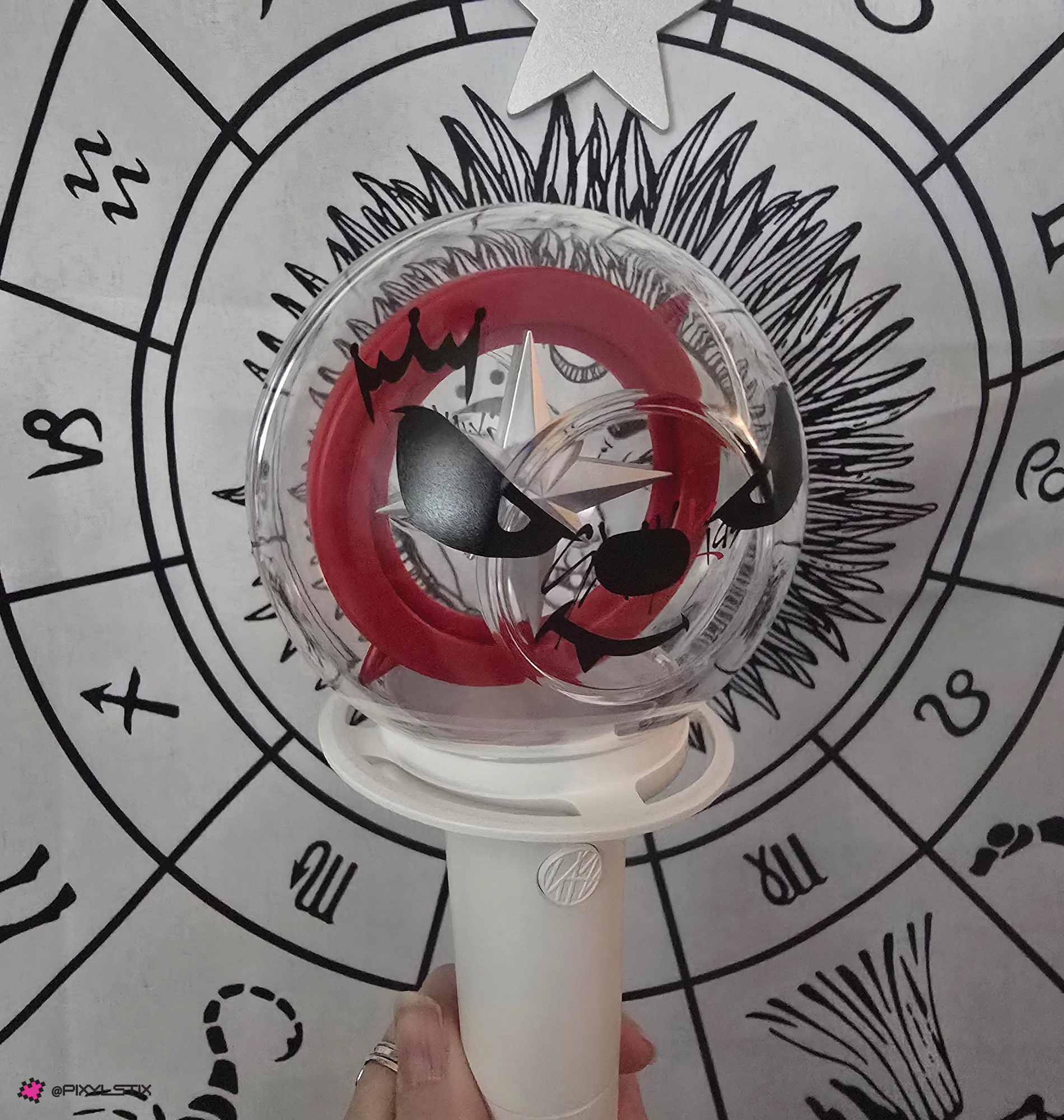 A stray kids lightstick with a face vinyl decal, held by a hand in front of a black-and-white zodiac wheel background.