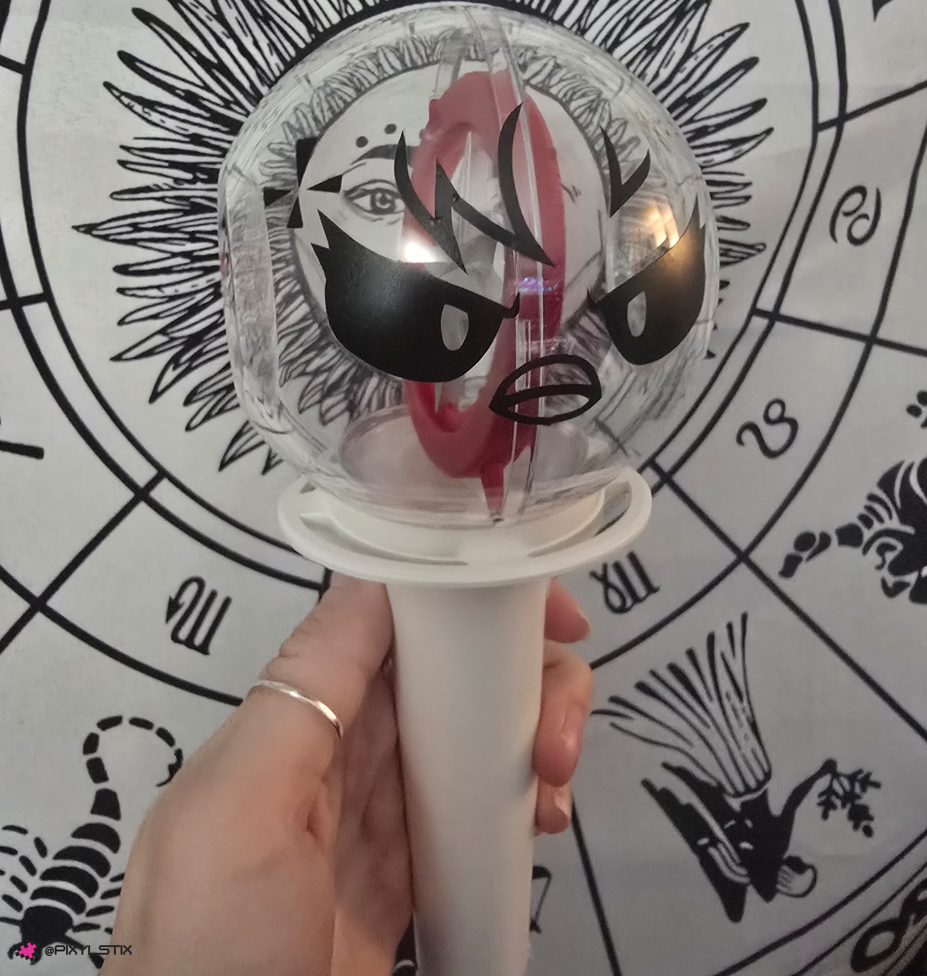 A stray kids lightstick with a face vinyl decal, held by a hand in front of a black-and-white zodiac wheel background.