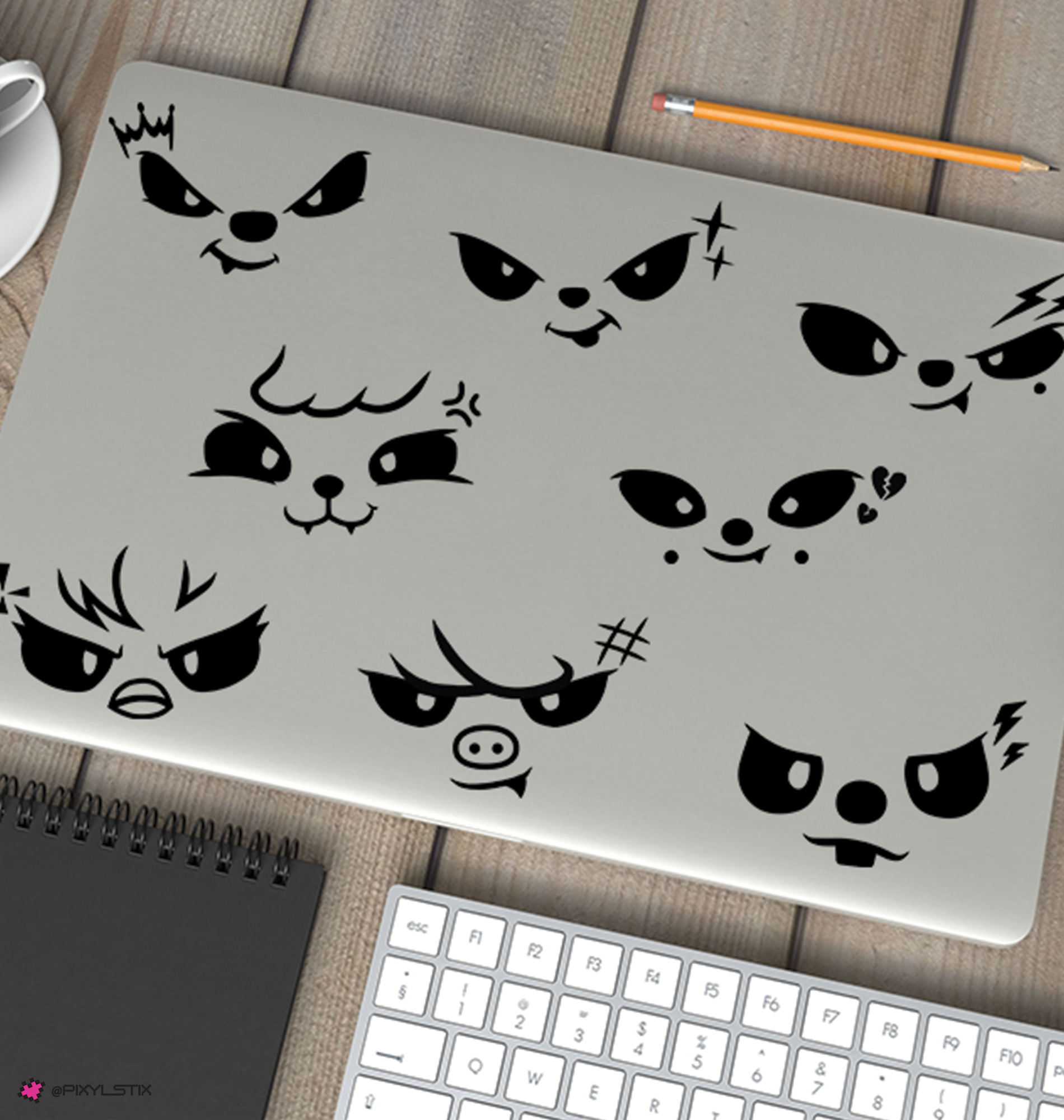 A silver laptop decorated with eight stray kids evil skzoo face decals, shown on a wooden desk with a keyboard, notebook, pencil, and coffee cup nearby.