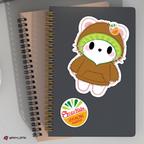 A spiral-bound notebook with a dark gray cover featuring a jiniret sticker wearing a brown hoodie and kiwi colored hood, plus a circular 'StravKids Hyunjin' fruit sticker. A beige notebook is partially visible behind it on a light gray background.