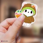 A hand holding a jiniret sticker wearing a brown hoodie and kiwi colored hood plus a circular fruit sticker.