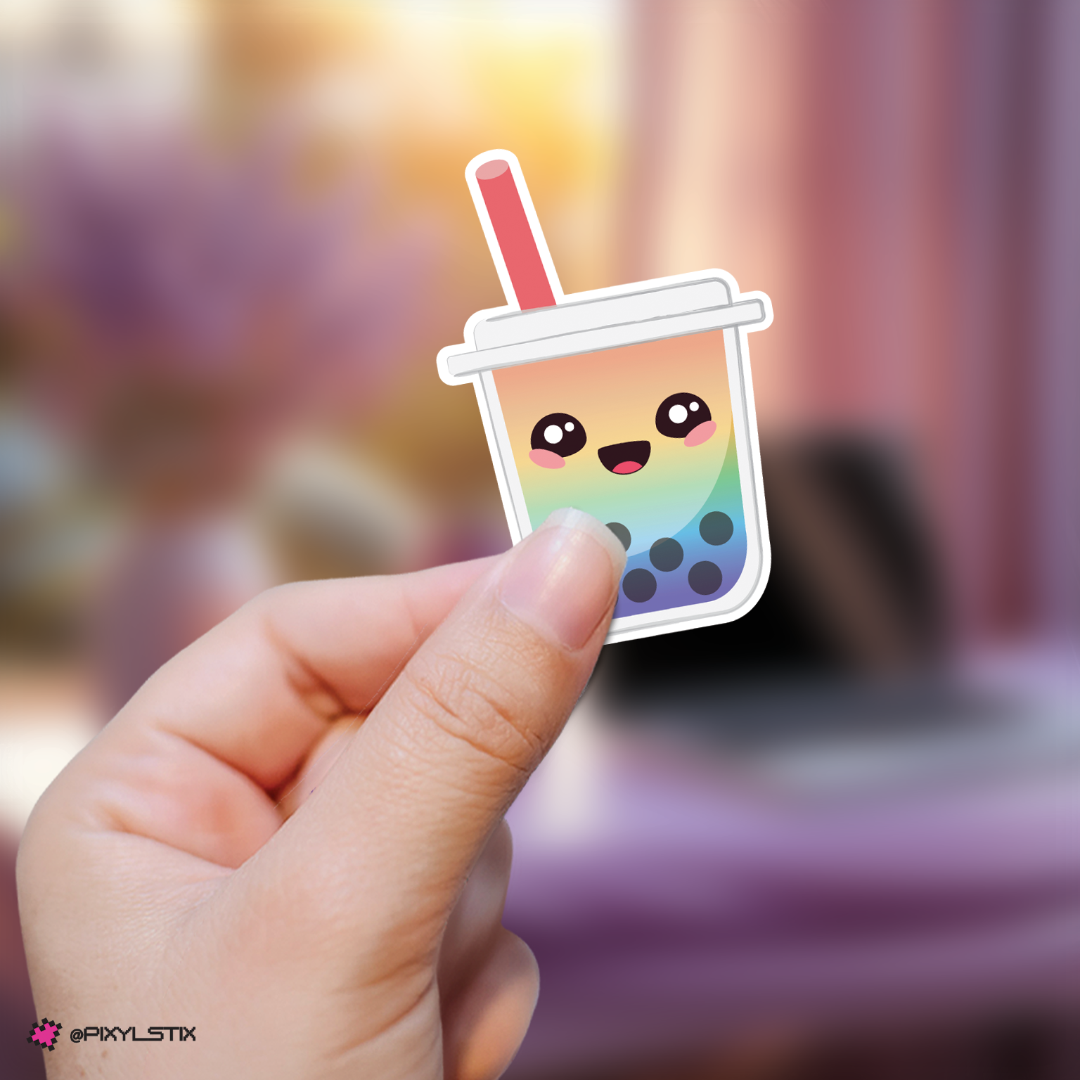 A hand holds a die-cut sticker shaped like a smiling boba tea cup with a rainbow gradient body, black bubble dots, and a pink straw, against a softly blurred indoor background.