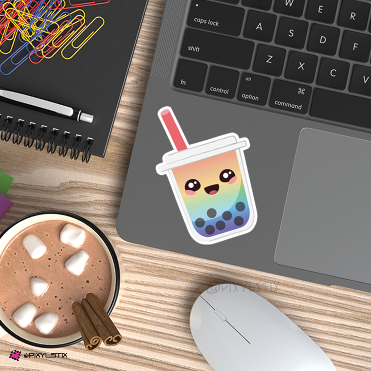 A cute cartoon boba tea cup sticker with a rainbow gradient and pink straw, shown applied to the palm rest of a gray laptop on a wooden desk next to a cup of hot chocolate and a computer mouse.
