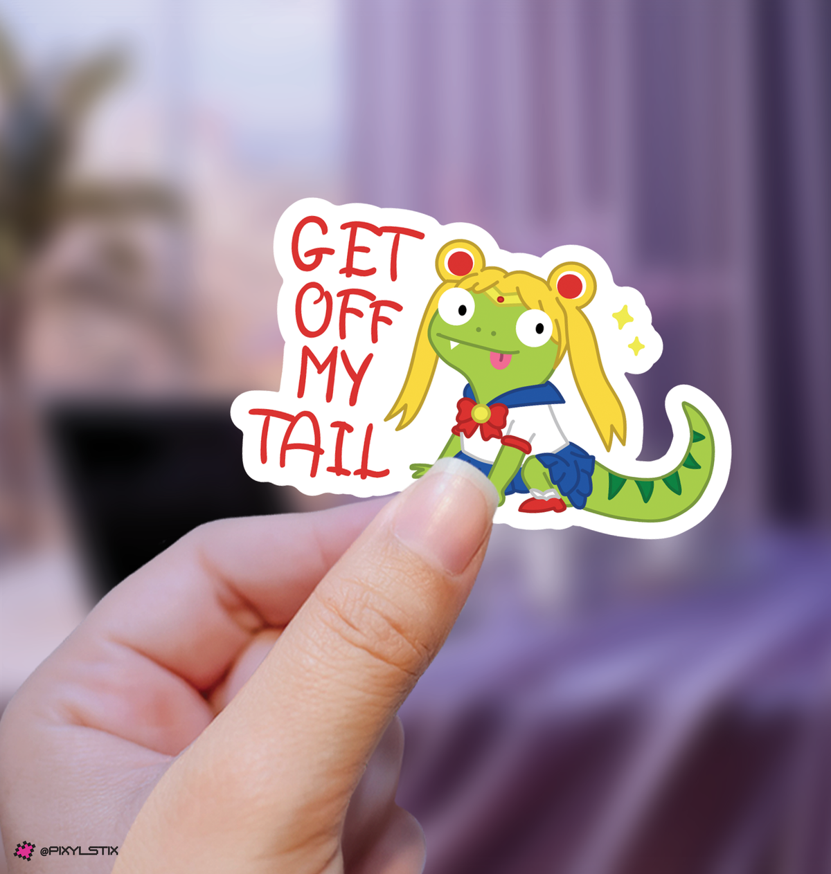 A hand holds a die-cut sticker featuring a cartoon green lizard character with yellow hair and the text 'GET OFF MY TAIL' in red, against a softly blurred purple indoor background.