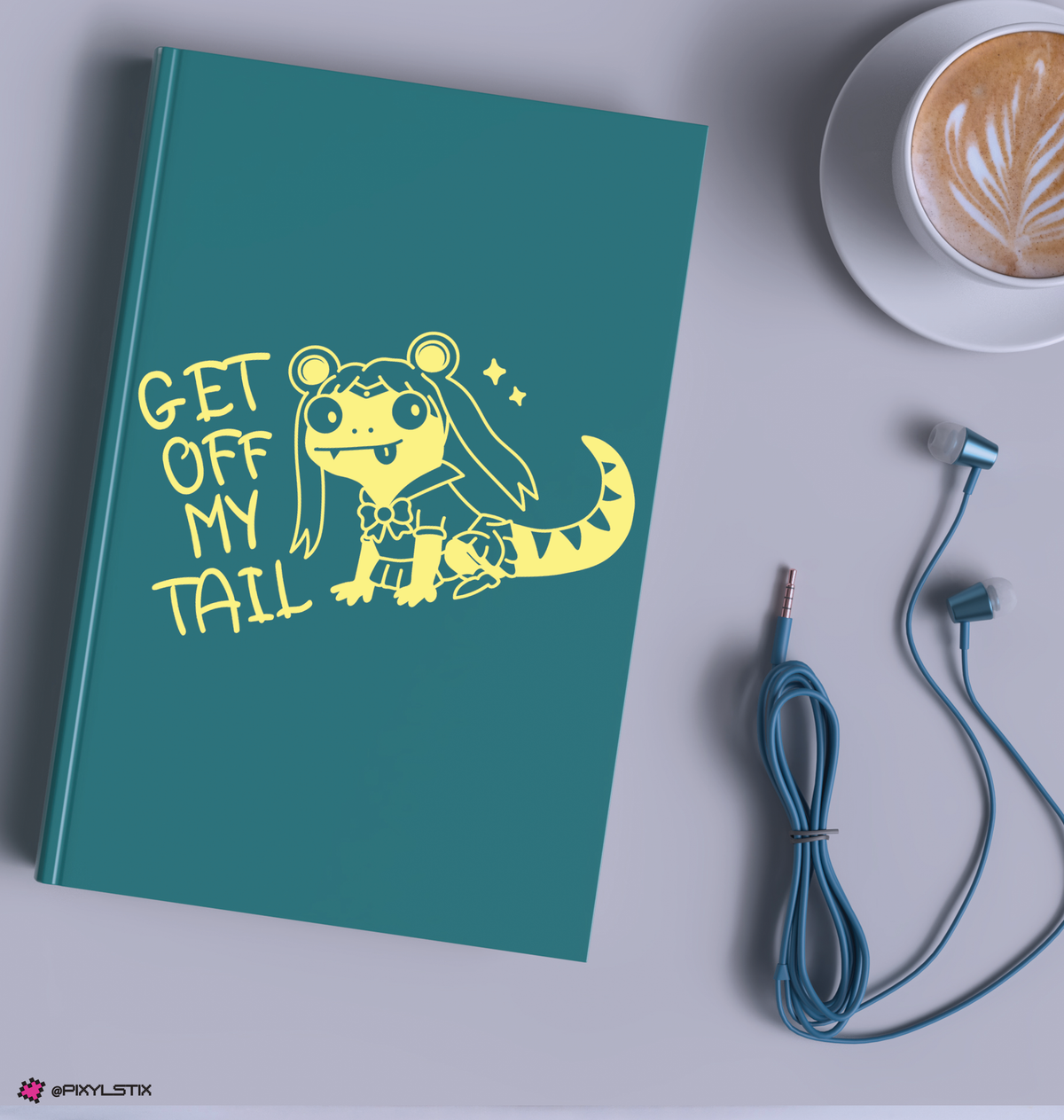 A teal notebook with a lizard cartoon character and the text 'GET OFF MY TAIL' is shown in a styled flat-lay on a light gray surface, alongside a cup of latte and blue wired earphones.