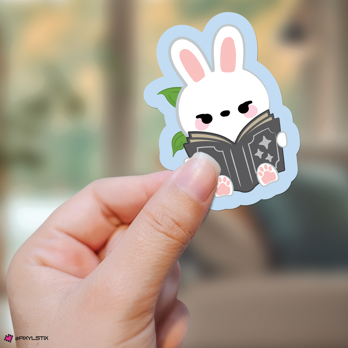 A hand holds a die-cut sticker featuring leebit reading a black book, with pink inner ears and paw pads, against a softly blurred indoor background.