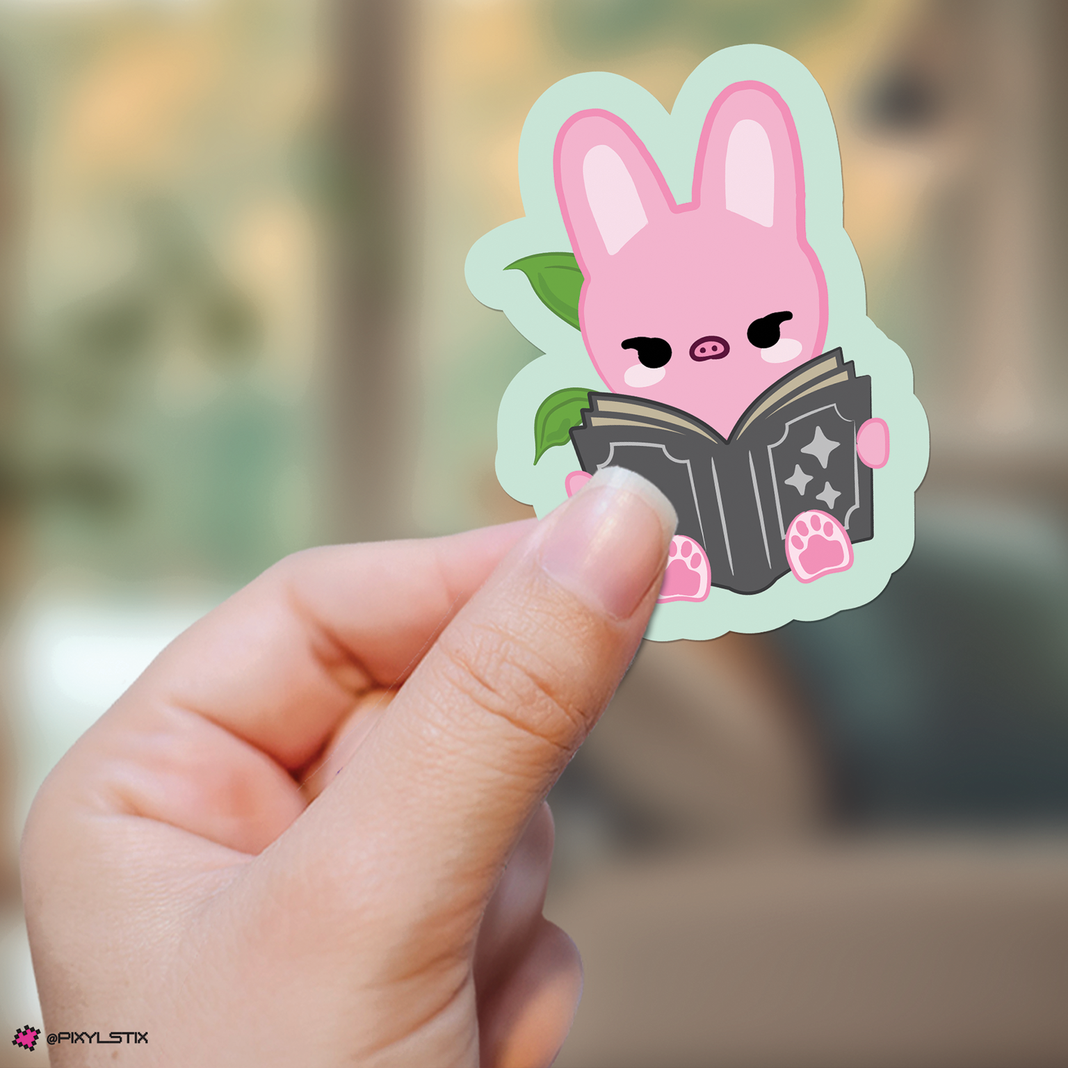 A hand holds a die-cut sticker featuring dwaekki reading a black book, with a light mint green outline and a blurred indoor background.