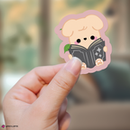 A hand holds a die-cut sticker featuring puppym reading a black book, with a soft pink outline and a blurred indoor background.