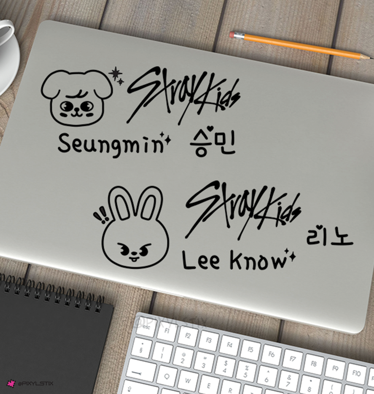 A silver laptop on a wooden desk with black vinyl decals of the Stray Kids members Seungmin and Lee Know, surrounded by a keyboard, notebook, pencil, and coffee cup.