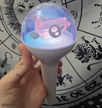 A hand holding a glowing white light stick with a translucent dome that glows blue and pink, featuring a miniteen decal. The background shows a black-and-white zodiac-themed print.