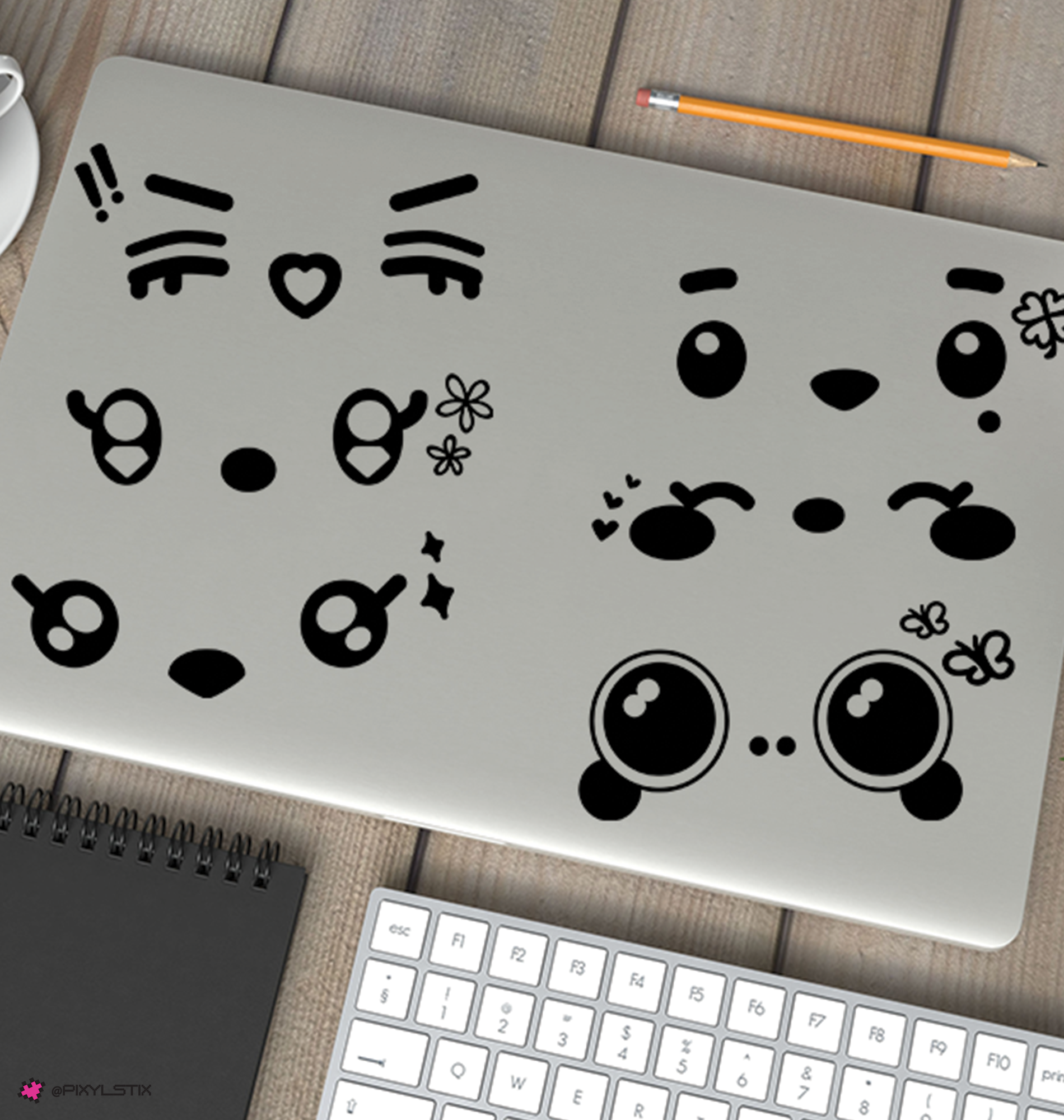 A silver laptop lid decorated with six black miniteen face decals, shown on a wooden desk with a keyboard, pencil, notebook, and coffee cup partially visible.
