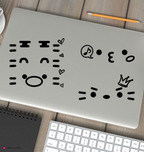 A silver laptop on a wooden desk with three black miniteed face decals on the lid. A keyboard, notebook, pencil, and partial cup are visible around it in a styled workspace setting.