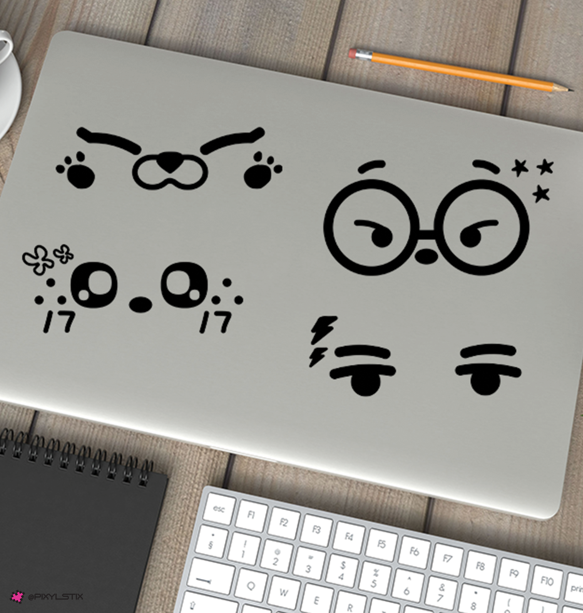 A silver laptop on a wooden desk with four black miniteed face decals on the lid. A keyboard, notebook, pencil, and partial cup are visible around it in a styled workspace setting.
