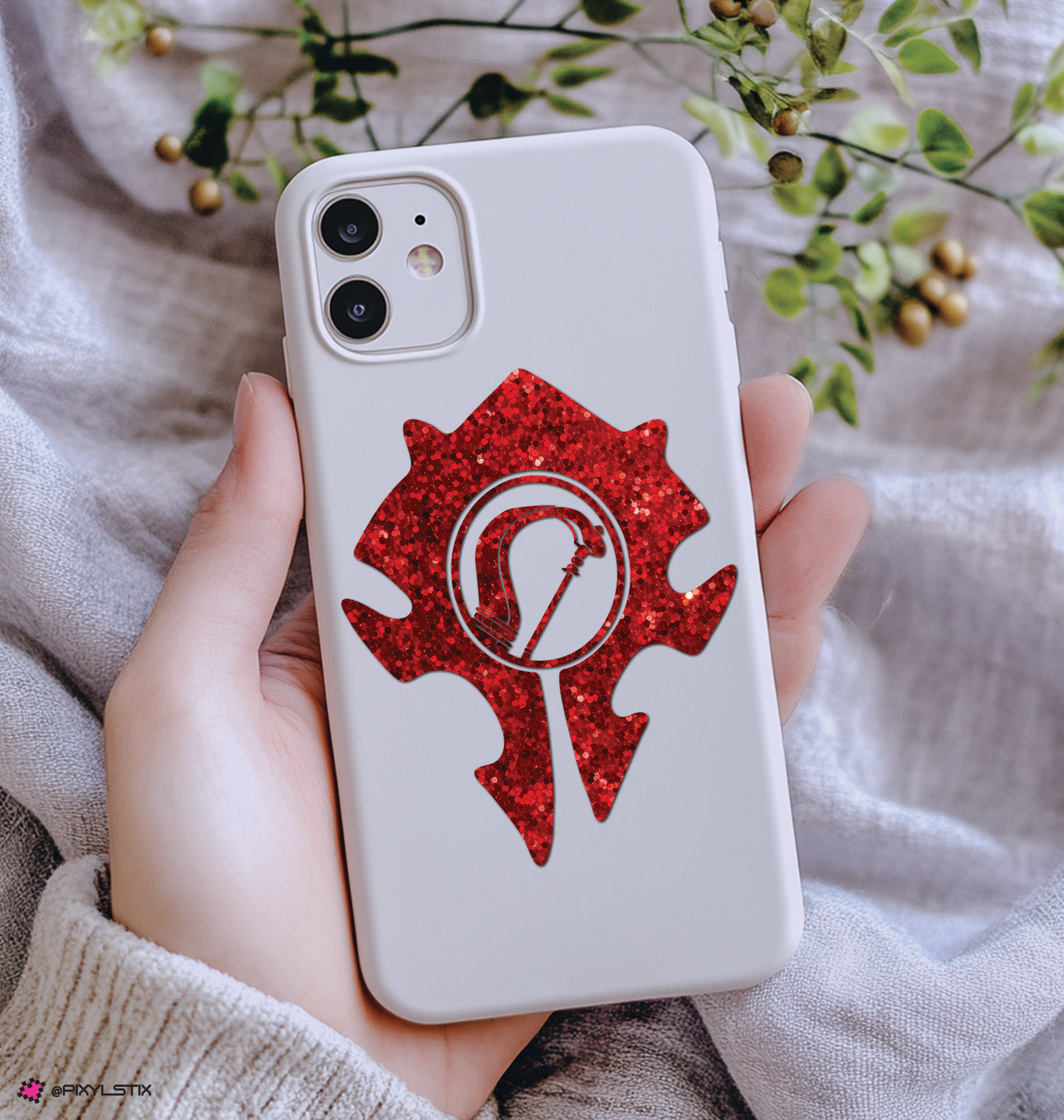 A hand holding a white phone case decorated with a red horde symbol featuring ahunter bow symbol in the center, set against a soft gray fabric background with green leaves.