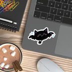 A die-cut vinyl sticker of a cute black bat with pink details and blue sparkles, shown applied to the palm rest of a gray laptop on a wooden desk next to a hot chocolate cup, mouse, and notebook.