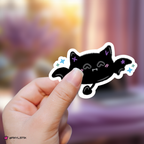 A hand holds a die-cut sticker of a black bat with purple and blue stars, against a blurred purple and pink background.
