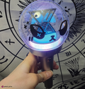 A hand holding a lightstick that glows with blue and purple with an xdinary monsters face decal on the surface. The background is a black and white astrological chart print.