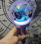 A hand holding a lightstick that glows with blue and purple with an xdinary monsters face decal on the surface. The background is a black and white astrological chart print.