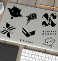 A laptop lid decorated with six black graphic stickers featuring stylized logos and the words 'Xdinary Heroes' and 'Villains', shown on a wooden desk with a keyboard, notebook, pencil, and cup partially visible.