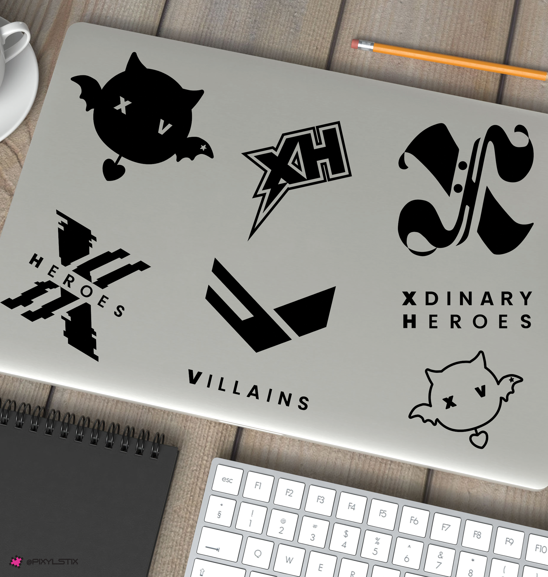 A laptop lid decorated with six black graphic stickers featuring stylized logos and the words 'Xdinary Heroes' and 'Villains', shown on a wooden desk with a keyboard, notebook, pencil, and cup partially visible.