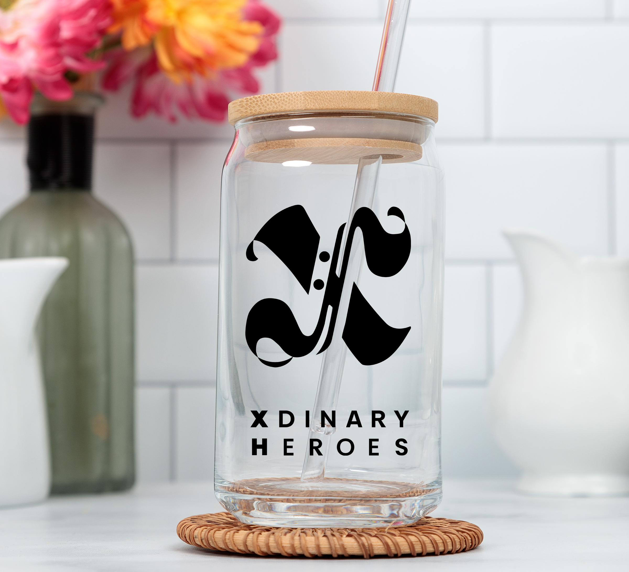 A clear glass tumbler with a bamboo lid and straw, featuring a black stylized 'XH' logo and the text 'XDINARY HEROES' on the front, set on a woven coaster in a kitchen-like setting.