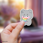 Hand holding a zeroni gunwook sticker with a rainbow heart against a blurred background