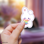 Hand holding a zeroni yujin sticker with a rainbow heart against a blurred background